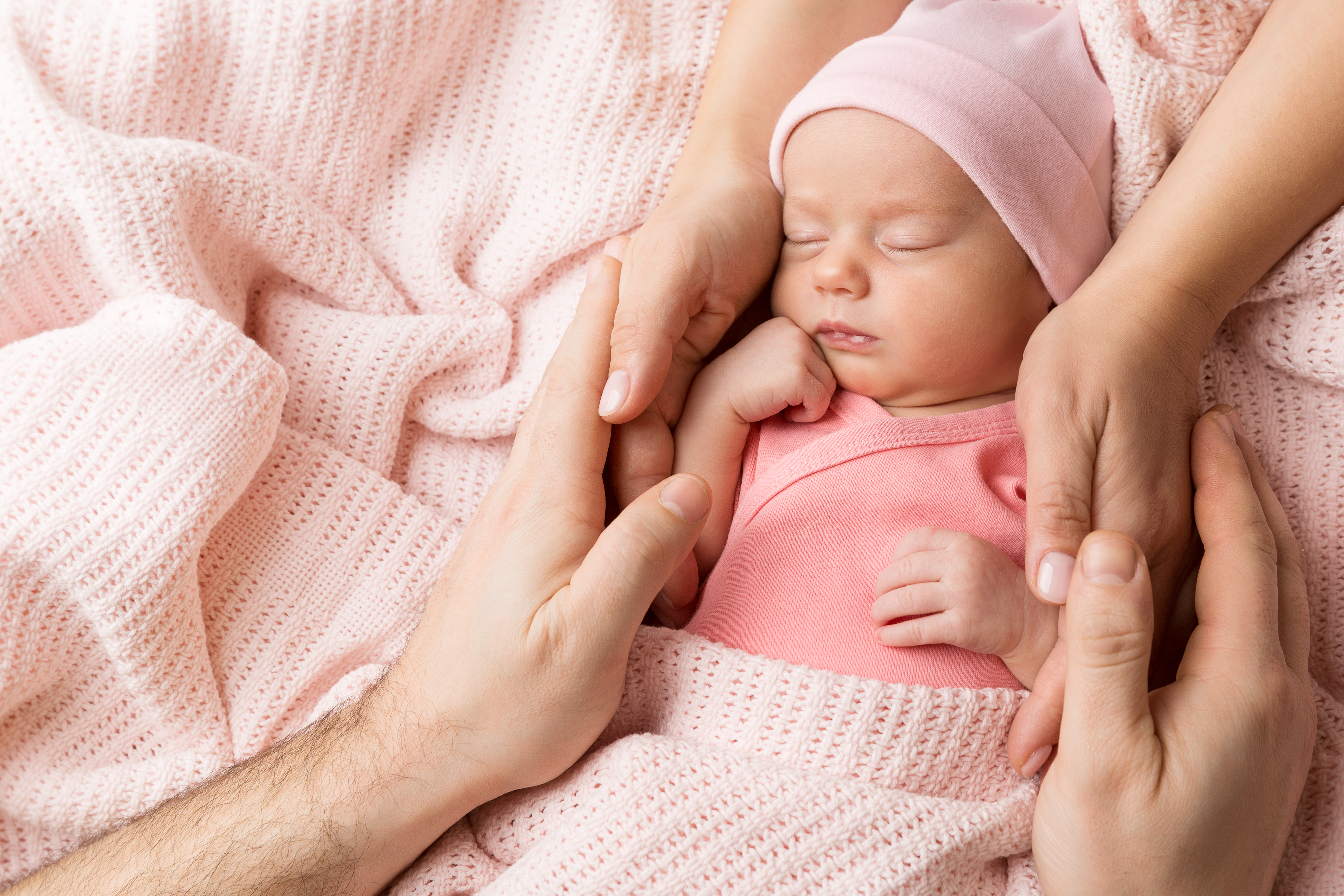 A baby wearing a pink outfit and hat lays on a pink blanket. Large, adult hands cradle the baby.